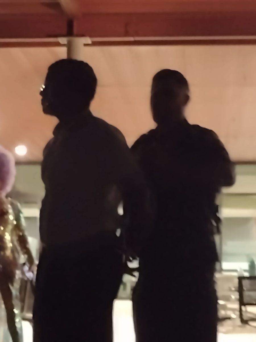 A student is being cuffed by a campus police officer as a drag performer stands in the background.
